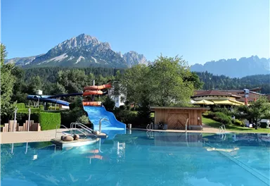 A beautiful pool with a water slide and a view of the mountains. Surrounded by green trees and sunny weather.