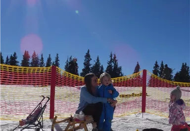 Eine Mutter sitzt mit ihrem Kind im Schnee und umarmt es. Im Hintergrund sind Bäume und ein sonniger Himmel zu sehen.