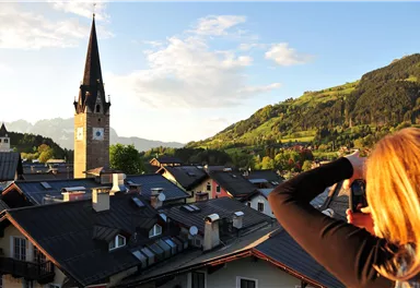 A woman is photographing the view of a picturesque village with a tall church tower. In the background, green hills and a clear sky can be seen.