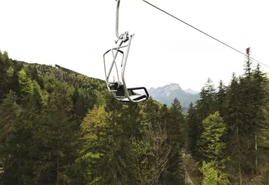 Eine Einsitzbahn schwebt über einen Wald mit Bergen im Hintergrund. Die Umgebung ist grün und bewaldet.