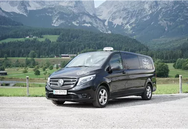 Ein schwarzer Van von Mercedes-Benz steht auf einer gravelartigen Fläche vor einer malerischen Berglandschaft. Im Hintergrund sind grüne Wiesen und hohe Berge sichtbar.
