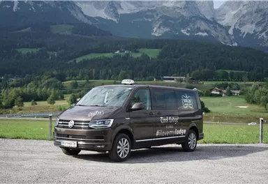 Ein brauner Volkswagen-Taxi-Van steht auf einem Parkplatz mit einer malerischen Berglandschaft im Hintergrund. Die Umgebung ist grün und hügelig unter einem klaren Himmel.