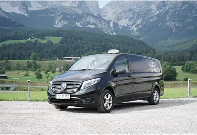 Ein schwarzer Van von Mercedes-Benz steht auf einer gravelartigen Fläche vor einer malerischen Berglandschaft. Im Hintergrund sind grüne Wiesen und hohe Berge sichtbar.