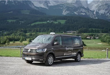 Ein brauner Volkswagen-Taxi-Van steht auf einem Parkplatz mit einer malerischen Berglandschaft im Hintergrund. Die Umgebung ist grün und hügelig unter einem klaren Himmel.
