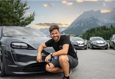 A man is posing smiling next to a modern car in a mountainous landscape. In the background, more cars and a beautiful sky can be seen.