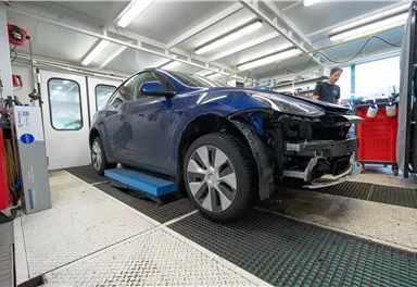 A blue car is in a workshop on a lift. In the background, a person is working on a car.