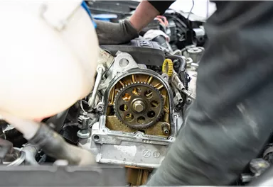 A mechanic is working on a car engine. Parts of the engine are visible, including a gear mechanism.