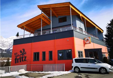 A modern building with an orange and gray design. It has a balcony and is located in a mountainous area.