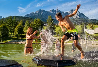 A group of children is playing in the water and splashing around. In the background, there are mountains and green trees visible.