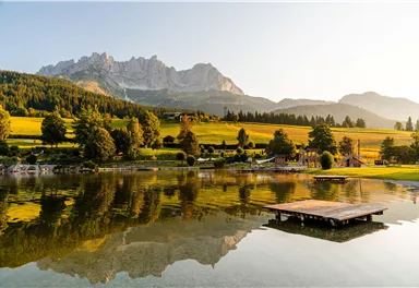 A picturesque lake with gentle hills and reflecting mountains in the background. The golden evening sun illuminates the landscape and creates a tranquil atmosphere.