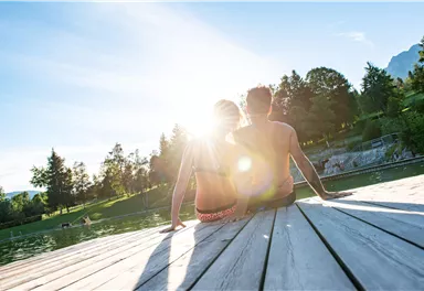 A couple is sitting on a wooden dock by the water, enjoying the sunset. The atmosphere is relaxed and romantic with a beautiful natural backdrop.
