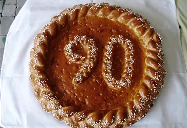 A large round cake with the number 90 on it, decorated with icing and sprinkles. The cake is placed on a white cloth.