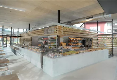 Ein modernes Café mit hellen, offenen Räumen und einer großen Theke, die verschiedene Lebensmittel anbietet. Die Einrichtung besteht aus Holz und bietet eine einladende Atmosphäre.