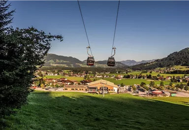 Eine Seilbahn, die über eine grüne Wiese schwebt, mit Blick auf ein kleines Dorf und die umliegenden Berge. Der Himmel ist klar und blau.