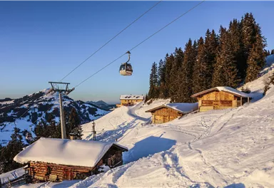 Ein malerisches Winterland mit schneebedeckten Hütten und einer Seilbahn. Hohe Tannenbäume umgeben die Szene, während die Sonne am klaren Himmel scheint.