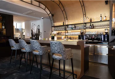 Eine elegante Bar mit stilvollen Stühlen und einer modernen Theke. Im Hintergrund sind Regale mit Getränken und Gläsern sichtbar.
