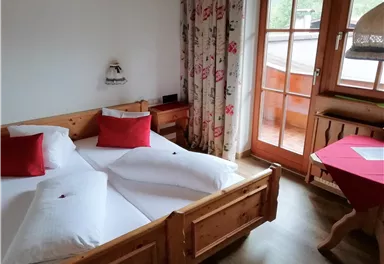 Ein gemütliches Schlafzimmer mit zwei Betten und roten Kissen. Der Raum hat einen Balkon mit Blick nach draußen und blumige Vorhänge.