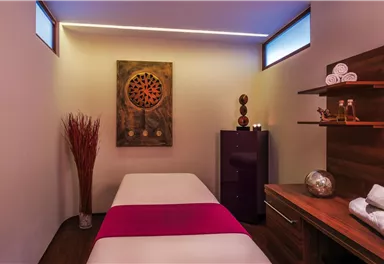 A relaxing treatment room with a massage table and soft lighting. The room contains decorative elements and towels that create a tranquil atmosphere.