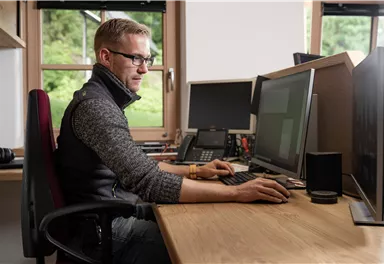 Ein Mann sitzt an einem Schreibtisch und arbeitet an einem Computer. Im Hintergrund sind weitere Bildschirme und Bürogeräte sichtbar.