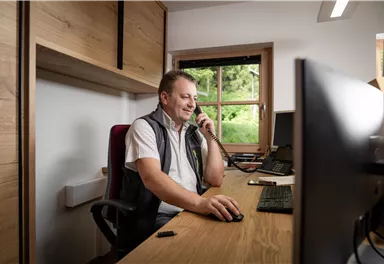 Ein Mann sitzt an einem Schreibtisch und telefoniert. Im Hintergrund ist ein Fenster mit Blick ins Grüne zu sehen.