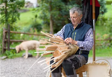 Ein Handwerker sitzt im Freien und flechtet mit Holzstücken. Im Hintergrund ist ein Hund zu sehen und die Natur ist grün und einladend.
