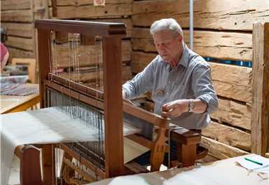 Ein älterer Mann arbeitet an einem Webstuhl in einer rustikalen Umgebung. Er konzentriert sich auf das Weben von Stoffen.