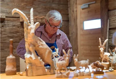 Ein Handwerker arbeitet konzentriert an Holzskulpturen von Tieren. Um ihn herum stehen verschiedene kunstvoll geschnitzte Figuren.