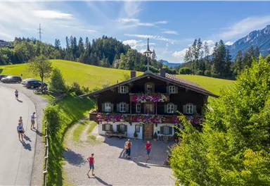 Ein malerisches alpenländisches Haus umgeben von grünen Wiesen und Bäumen. Vor dem Haus sind Menschen und Autos zu sehen, mit Bergen im Hintergrund.