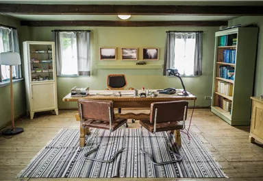 Ein gemütliches Büro mit einem großen Holzschreibtisch und zwei Stühlen. Die Wände sind grün gestrichen, und es gibt Regale und Fenster mit Vorhängen.