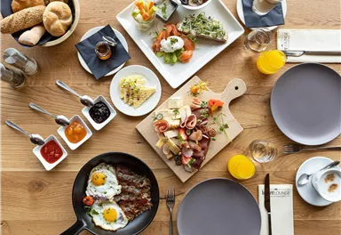 Ein reichhaltiges Frühstück auf einem Holztisch, bestehend aus Rühreiern, Aufschnitt, Brot und frischem Obst. Dazu gibt es verschiedene Saucen in kleinen Schalen und Getränke.