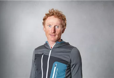 Ein Mann mit roten Haaren trägt eine sportliche Jacke. Er steht vor einem neutralen Hintergrund und schaut direkt in die Kamera.