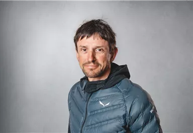 Ein Mann mit kurzer Frisur trägt eine blaue Daunenjacke. Er steht vor einem neutralen, grauen Hintergrund.