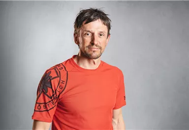 Ein Mann trägt ein rotes T-Shirt mit einem Emblem. Der Hintergrund ist grau und neutral.