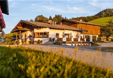 Ein traditionelles Gebäude im ländlichen Stil, umgeben von Wiesen und Wäldern. Die Terrasse ist einladend mit Tischen und Sonnenschirmen.