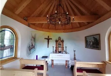 Eine kleine Kapelle mit einem Holzdach und einer altarähnlichen Einrichtung. An den Wänden sind religiöse Bilder und ein Kreuz zu sehen.