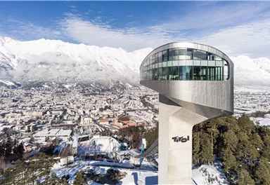 A modern observation tower with unique architecture stands majestically in a snow-covered landscape. In the background, impressive mountains and an urban landscape stretch out.
