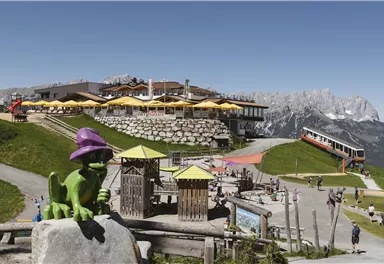 Eine Berglandschaft mit einem Restaurant und sonnigen Terrassen. Im Vordergrund steht eine grüne Figur mit einem lila Hut und Spielgeräte für Kinder.