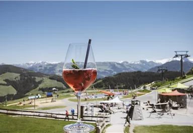 Ein erfrischendes Getränk in einem Glas steht im Vordergrund. Im Hintergrund sind eine wunderschöne Berglandschaft und eine Freizeitarea zu sehen.