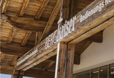 Ein rustikales Holzgebäude mit dem Schild „Bergkäserei“ und der Aufschrift „Bedienungsrestaurant“. Im Vordergrund sind bunte Blumen zu sehen.