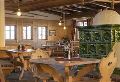 Ein gemütliches Restaurant mit Holzmöbeln und einem grünen Ofen. Die Wände sind aus Holz, und große Fenster sorgen für viel Licht.
