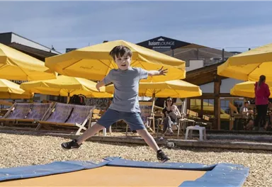 Ein Junge springt fröhlich auf einem Trampolin. Im Hintergrund gibt es sonnige Liegestühle und Sonnenschirme.