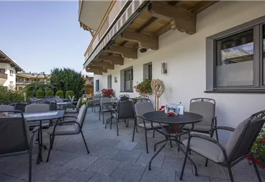 Eine gemütliche Terrasse mit Tischen und Stühlen. Im Hintergrund sind Pflanzen und ein klarer Himmel zu sehen.