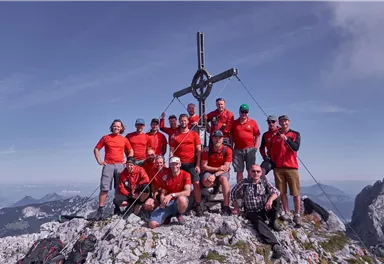 A group of ten people is standing on a mountain peak, surrounded by rocks and a clear sky. They are wearing red T-shirts and are happily posing in front of a cross.