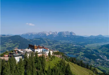 Eine malerische Berglandschaft mit einer Berghütte im Vordergrund. Im Hintergrund erheben sich majestätische Berge unter einem klaren blauen Himmel.