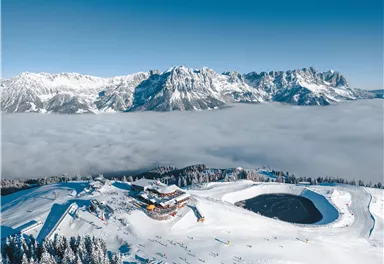 Eine schneebedeckte Berglandschaft mit majestätischen Bergen im Hintergrund. In der Mitte befindet sich eine Hütte, umgeben von Wolken und Skigebieten.