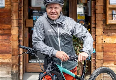 Ein Mann in einem grauen Regenjacke steht neben einem Mountainbike. Im Hintergrund ist eine Holzhütte zu sehen.