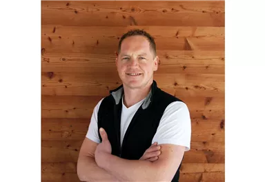 Ein lächelnder Mann steht mit verschränkten Armen vor einer Holzwand. Er trägt ein weißes T-Shirt und eine schwarze Weste.