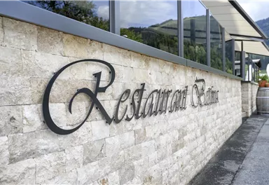 An elegant metal restaurant sign on a stone wall. In the background, windows and the surrounding nature are visible.