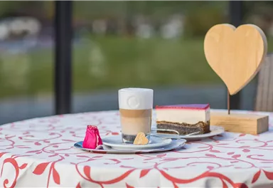 A decorative table with a cup of coffee and a piece of cake. In the background, a wooden heart stands as a decorative element.