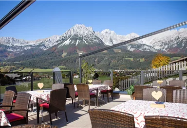 A cozy outdoor area with rattan-like chairs and tables. In the background, majestic mountains rise under a clear blue sky.
